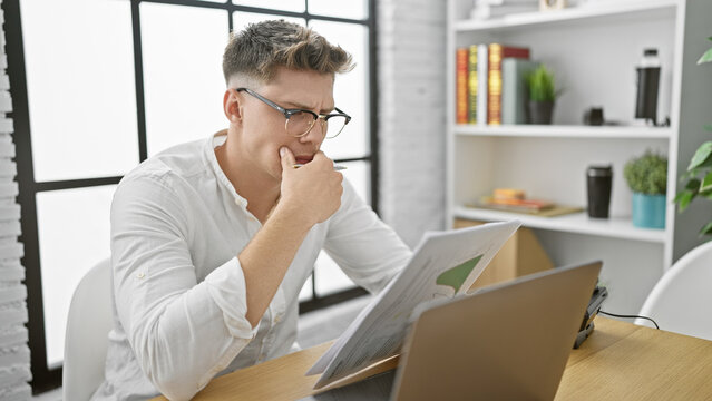 Handsome, bearded, young caucasian man, in glasses, seriously engrossed in reading a business document inside his office, a picture of focused professional in work mode.