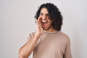 Fototapeta na wymiar Hispanic man with curly hair standing over white background shouting and screaming loud to side with hand on mouth. communication concept.