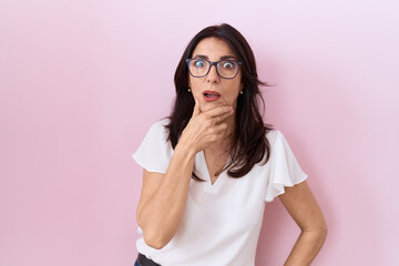 Middle age hispanic woman wearing casual white t shirt and glasses looking fascinated with disbelief, surprise and amazed expression with hands on chin