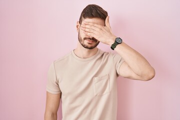 Hispanic man with beard standing over pink background covering eyes with hand, looking serious and...