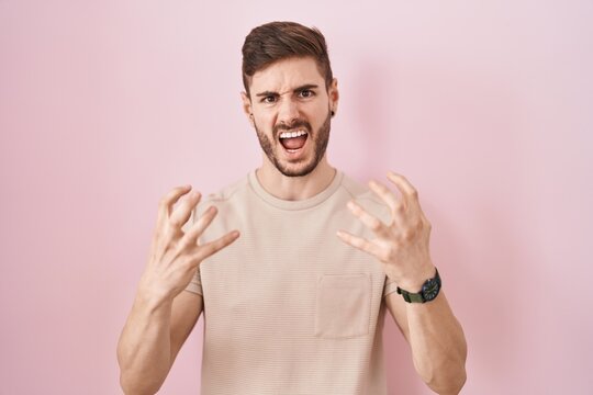 Hispanic man with beard standing over pink background crazy and mad shouting and yelling with aggressive expression and arms raised. frustration concept.
