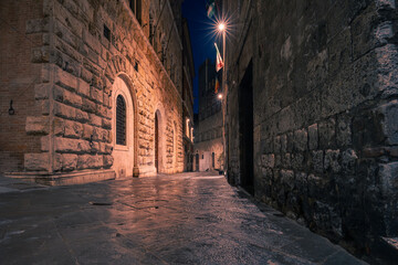 Dark narrow empty medieval street in the historical center of at night with rare lights, Siena,...