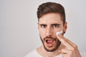 Hispanic man with beard using moisturizer facial cream afraid and shocked with surprise and amazed expression, fear and excited face.