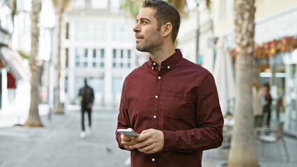 A bearded young hispanic man in a burgundy shirt looks thoughtfully while holding a smartphone on a...