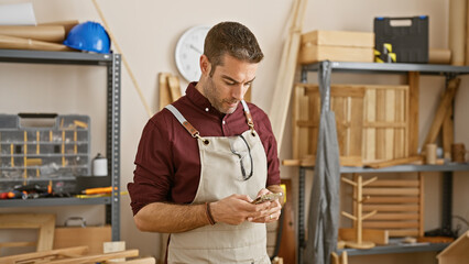 Handsome hispanic man with beard in workshop using smartphone, tools on background.