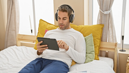 Relaxed man with headphones using tablet on bed in a bright, cozy bedroom