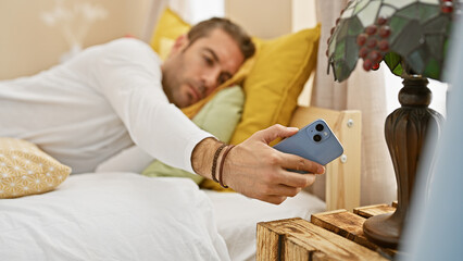 A young hispanic man with a beard lies in bed using his smartphone in a cozy home setting.