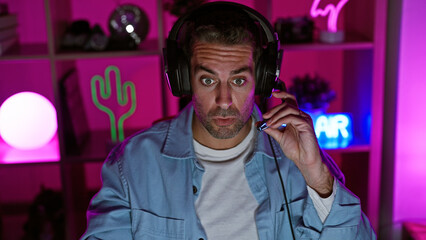 A handsome young hispanic man with a beard wearing headphones in a dark gaming room highlighted with pink neon lights.