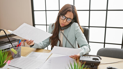 Stunning young hispanic woman, an emotive employee, engrossed in reading business documents, chats...