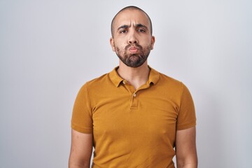 Hispanic man with beard standing over white background puffing cheeks with funny face. mouth...
