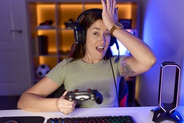 Beautiful brunette woman playing video games wearing headphones surprised with hand on head for...