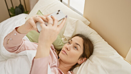 A young woman with brunette hair and blue eyes lies in bed, using her smartphone in a cozy, well-lit bedroom.