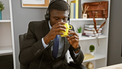A professional african american man sips coffee while wearing headphones at his office desk,...