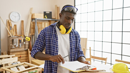 African american man working attentively in a carpentry workshop with tools, wood, and safety...