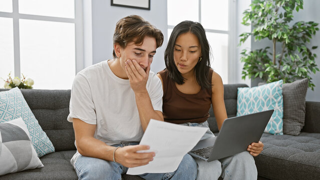Interracial couple analyzes documents in a modern living room with a laptop, expressing concern and teamwork.