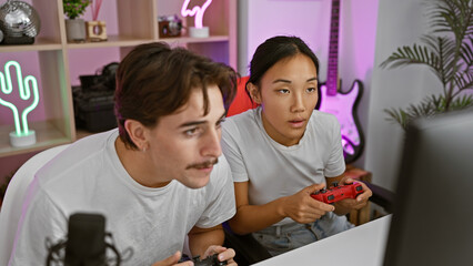 A man and a woman engaged in gaming, sitting indoors with neon lights illuminating their focused...