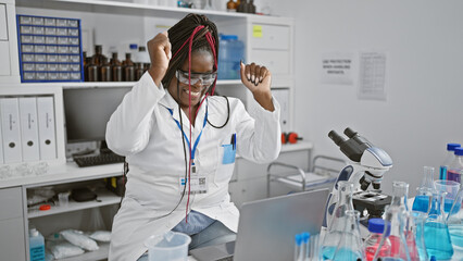 Thrilled african american female scientist, rocking her braids & glasses, strikes a win-celebration gesture at lab while exploring medical wonders on laptop!