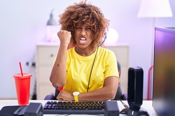 Young hispanic woman with curly hair playing video games wearing headphones angry and mad raising fist frustrated and furious while shouting with anger. rage and aggressive concept.