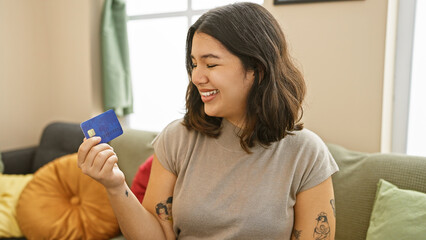 Smiling young hispanic woman holds a credit card in a cozy living room, expressing financial...
