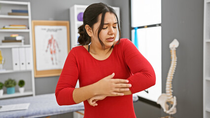 A young hispanic woman in pain holding her arm inside a rehab clinic room with anatomical posters.