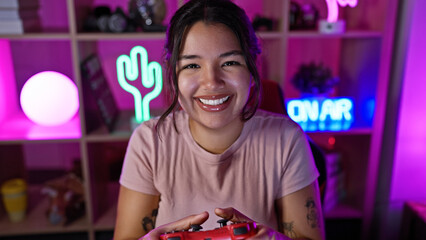 A smiling young hispanic woman holding a controller in a neon-lit gaming room at night.