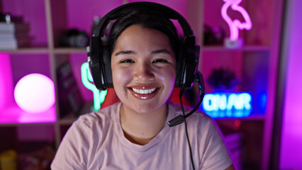 A smiling hispanic woman with headphones in a colorful gaming room at night.