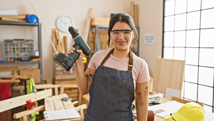 A smiling young woman in a workshop holding a drill portrays skilled carpentry indoors.