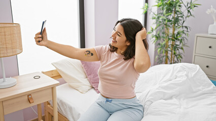 Smiling young hispanic woman taking a selfie in her well-lit, cozy bedroom, adding a touch of lifestyle and positivity.