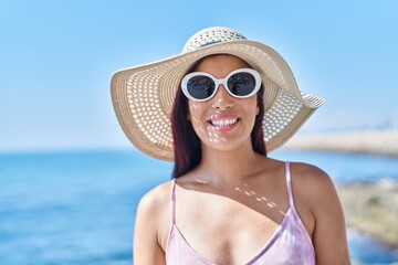 Young beautiful hispanic woman tourist smiling confident wearing hat and sunglasses at seaside