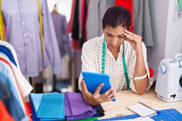 Young beautiful hispanic woman tailor stressed using touchpad at tailor shop