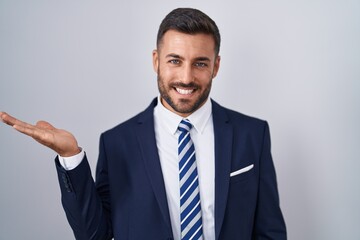 Handsome hispanic man wearing suit and tie smiling cheerful presenting and pointing with palm of hand looking at the camera.