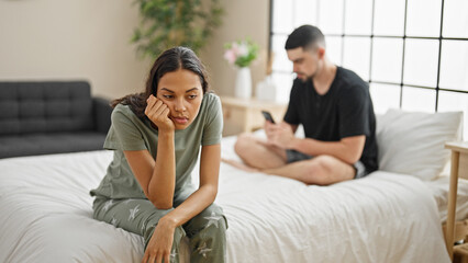 Beautiful couple disagreeing in their bedroom, sitting on the bed, expressing conflict while using...