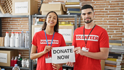 Man and woman volunteers, standing with paper herald, urging 'donate now' at charity center,...