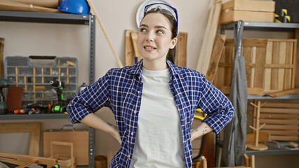 Confident young woman in a plaid shirt stands hands-on-hips in a well-organized carpentry workshop, embodying a professional woodworker's poise.