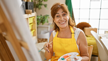 Mature woman painting in an art studio, smiling joyfully at the camera, wearing a yellow apron.