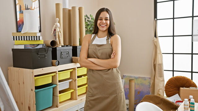 Attractive young hispanic woman artist standing with a confident, relaxed smile, arms crossed in her indoor art studio. a beautiful portrait of her juggling creativity, education and hobby.