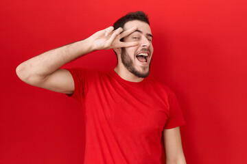 Young hispanic man wearing casual red t shirt doing peace symbol with fingers over face, smiling cheerful showing victory