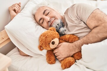 Middle age grey-haired man hugging teddy bear lying on bed sleeping at bedroom