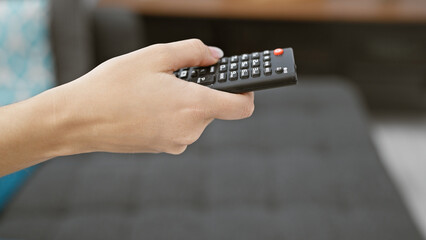 Close-up of a person's hand holding a black television remote control in a blurred indoor living...