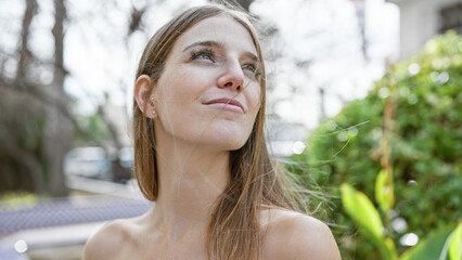 Young, beautiful, caucasian woman gazing upward in a lush outdoor garden setting, embodying a sense of contemplation.