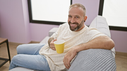 Captivating snapshot of a smiling young man, comfortably sitting on his living room sofa, savoring a hot coffee drink at home, radiating positivity and confidence.