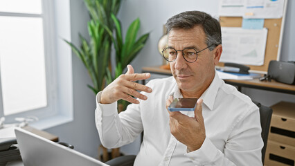 Mature businessman in glasses uses voice command on smartphone in modern office.