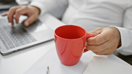 A middle-aged man in an office holding a red coffee mug while using a laptop, depicting a...