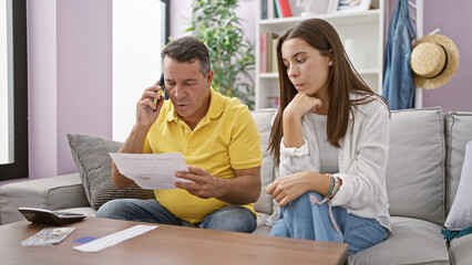Hispanic father and daughter reading receipt together on their home sofa, serious financial conversation via smartphone
