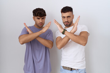 Homosexual gay couple standing over white background rejection expression crossing arms doing...