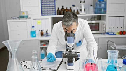 Attractive grey-haired young hispanic man, a dedicated scientist, working intently on a medical experiment in the lab, safety gloves on, peering through a microscope, deep in concentration.