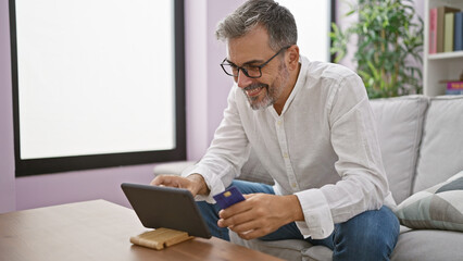 Young hispanic grey-haired man using touchpad and credit card sitting on sofa smiling at home