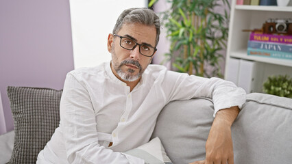 Troubled young hispanic man with grey-haired beard, wearing glasses, reveals a serious expression while sitting on living room sofa, indoors at home. a worrying sign of possible distress.