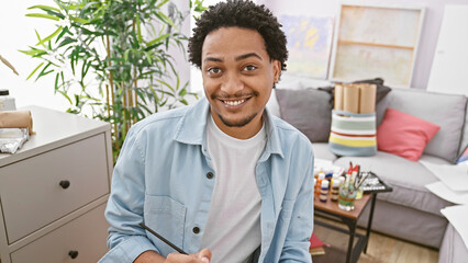 Portrait of a smiling young adult man with curly hair, casually dressed, sitting in front of a modern indoor studio setting.