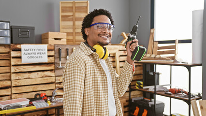 Handsome man with drill at indoor workshop wearing safety goggles and headphones.
